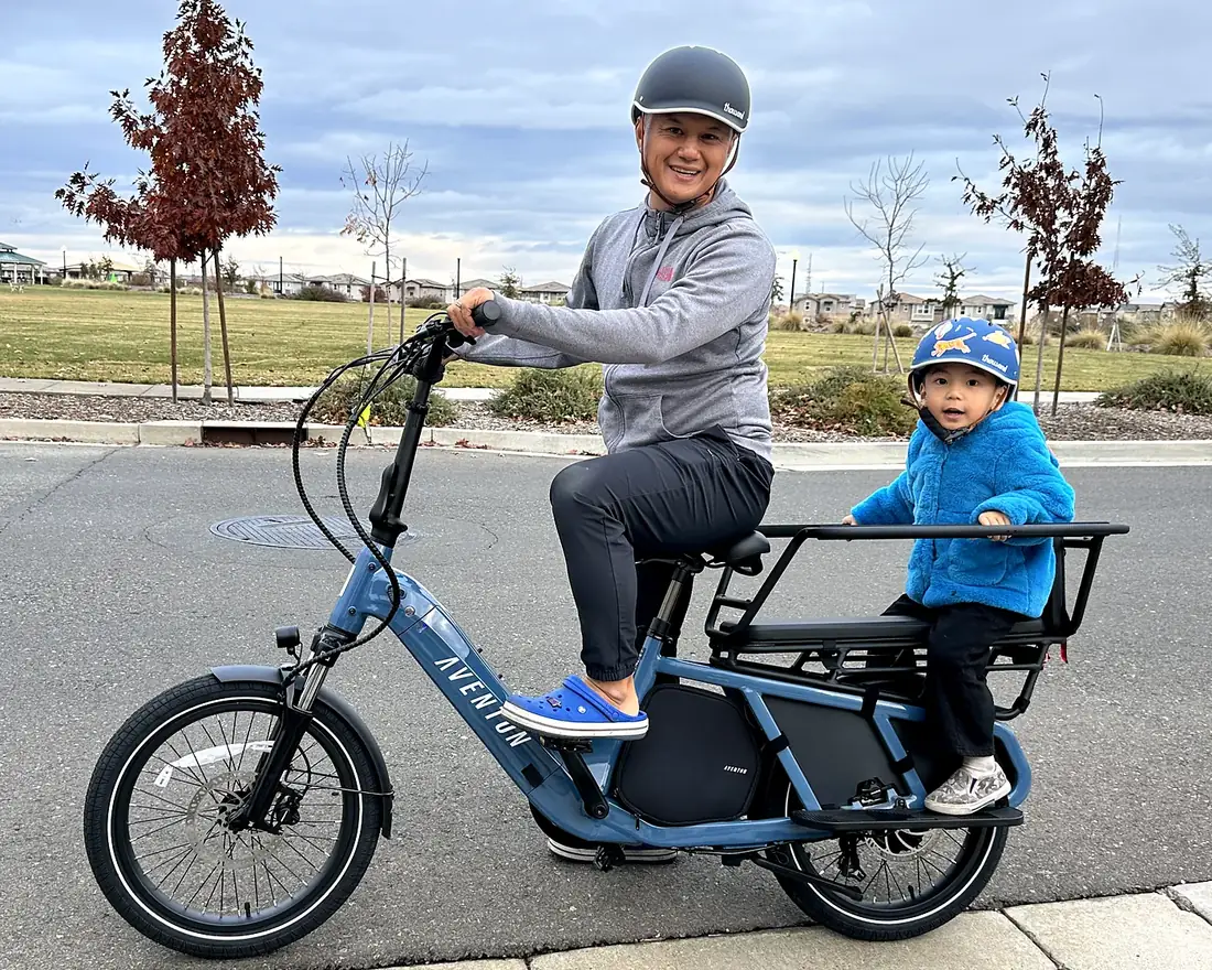 Marvin smiling on a blue cargo e-bike, one of his kids in a helmet on the back seat, on a street next to a park in Sacramento