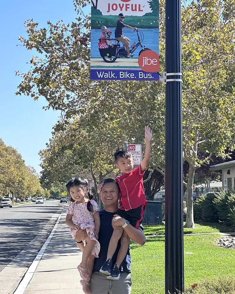 Marvin holding his son and daughter under a Walk. Bike. Bus. street banner that shows him riding a cargo bike with two of his kids