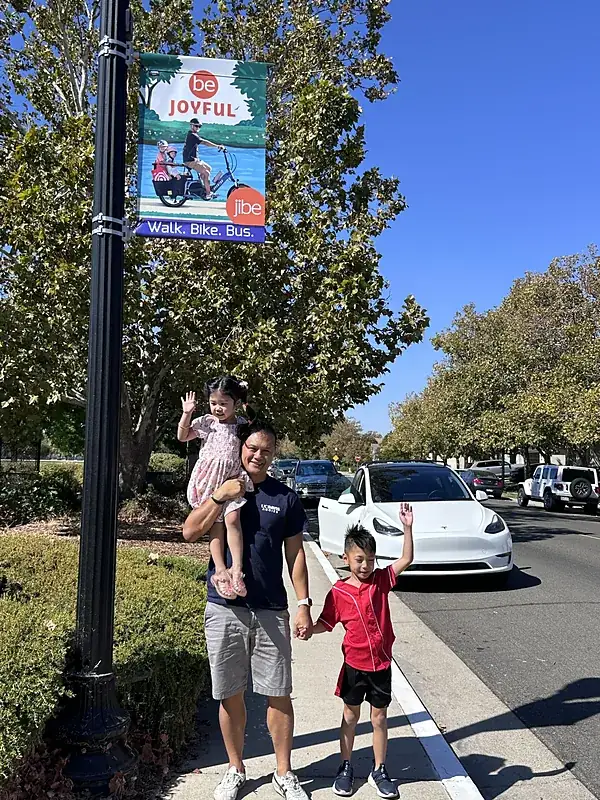 Marvin holding his daughter and his son's hand under another Walk. Bike. Bus. banner on a tree-lined street