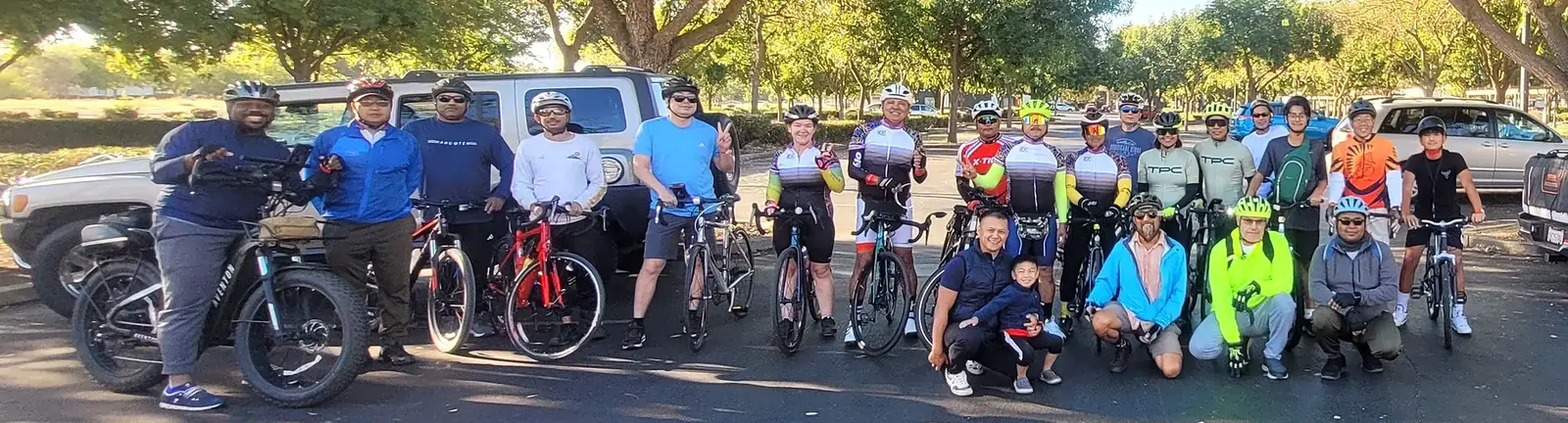 Marvin kneeling at the front of a group of about 25 riders with road bikes and e-bikes in a tree-lined parking lot before a community ride