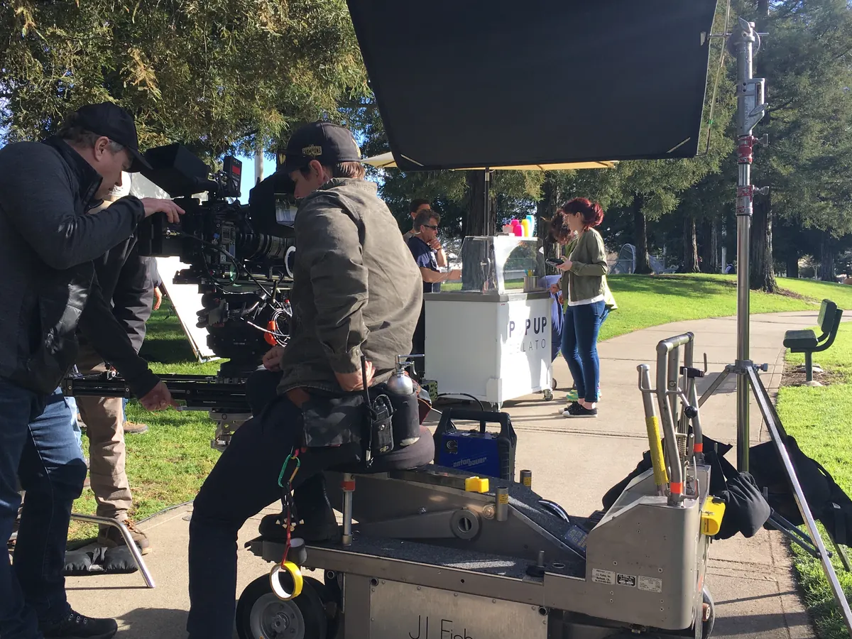 A film crew with a camera dolly in a park, a Pop Up Gelato cart set up behind them as part of the scene