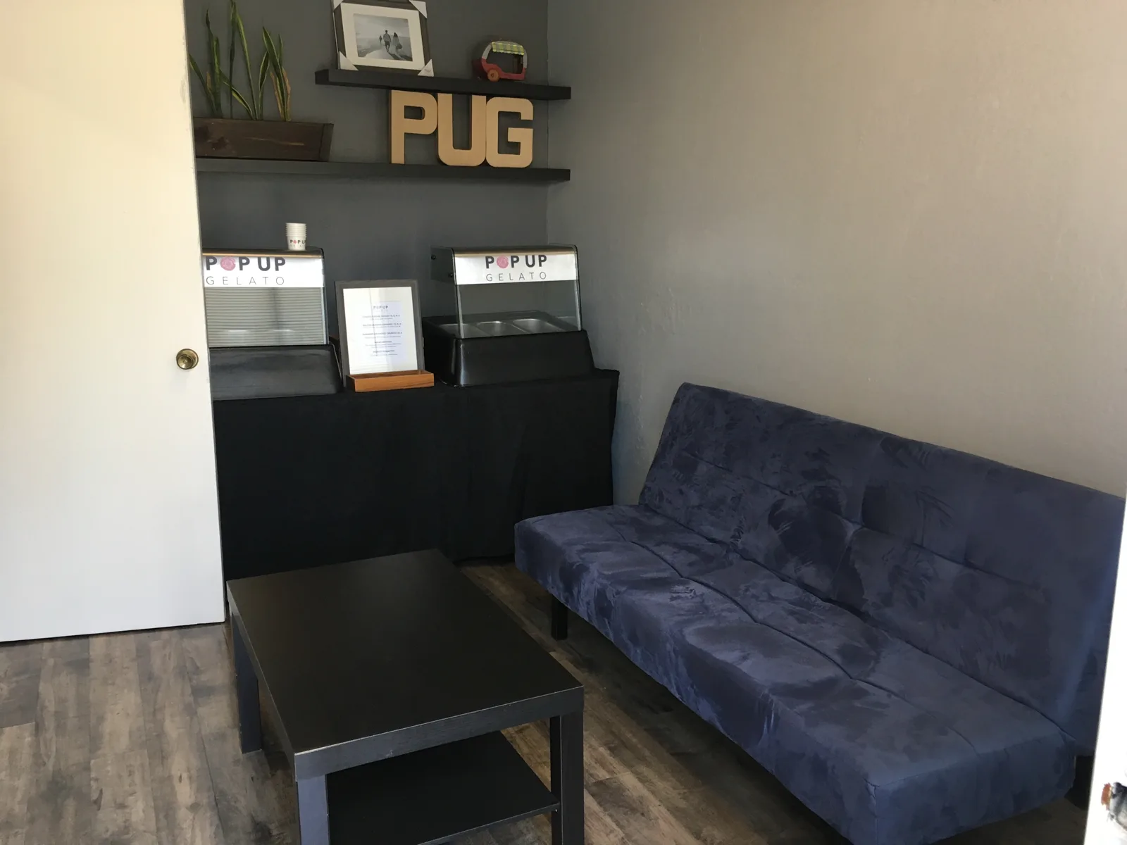 The Fremont tasting room: gray walls, floating shelves with wooden PUG letters, two branded stations on a black-draped table, a navy futon and dark coffee table on new wood-look flooring