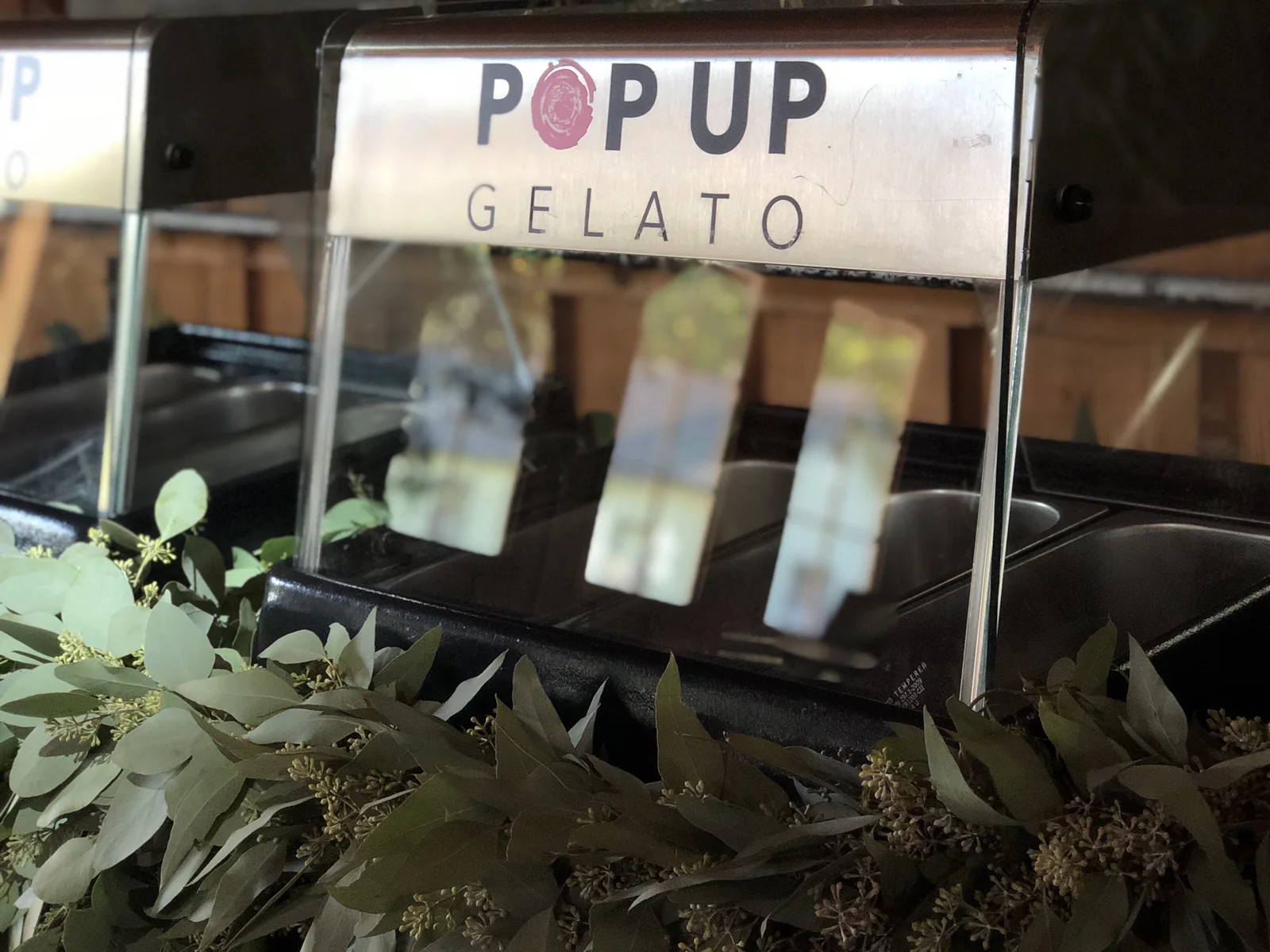 A Pop Up Gelato tabletop station with a brushed-steel sign and a eucalyptus garland along its base, set up at an outdoor event